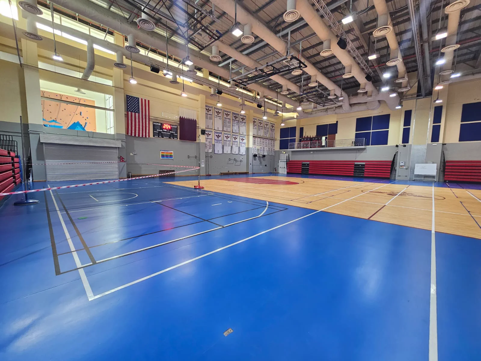 Multi-court school gymnasium in Doha with blue synthetic sports flooring, navy acoustic wall panels and folded red bleachers