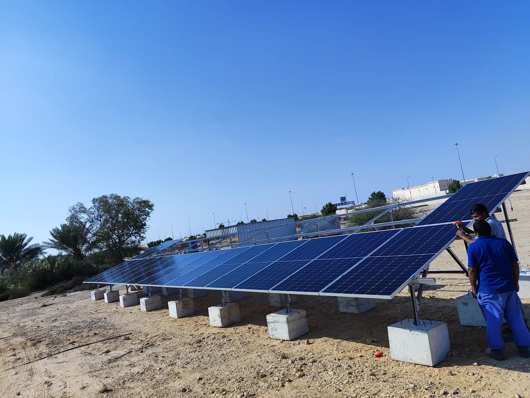 Ground-mounted photovoltaic array on concrete plinths powering a solar water pump, being cleaned by a technician in Qatar